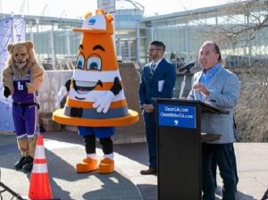 Man giving a speech at a podium with a person wearing a traffic cone costume and a lion costume.