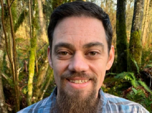 Man with a beard standing in the forest.